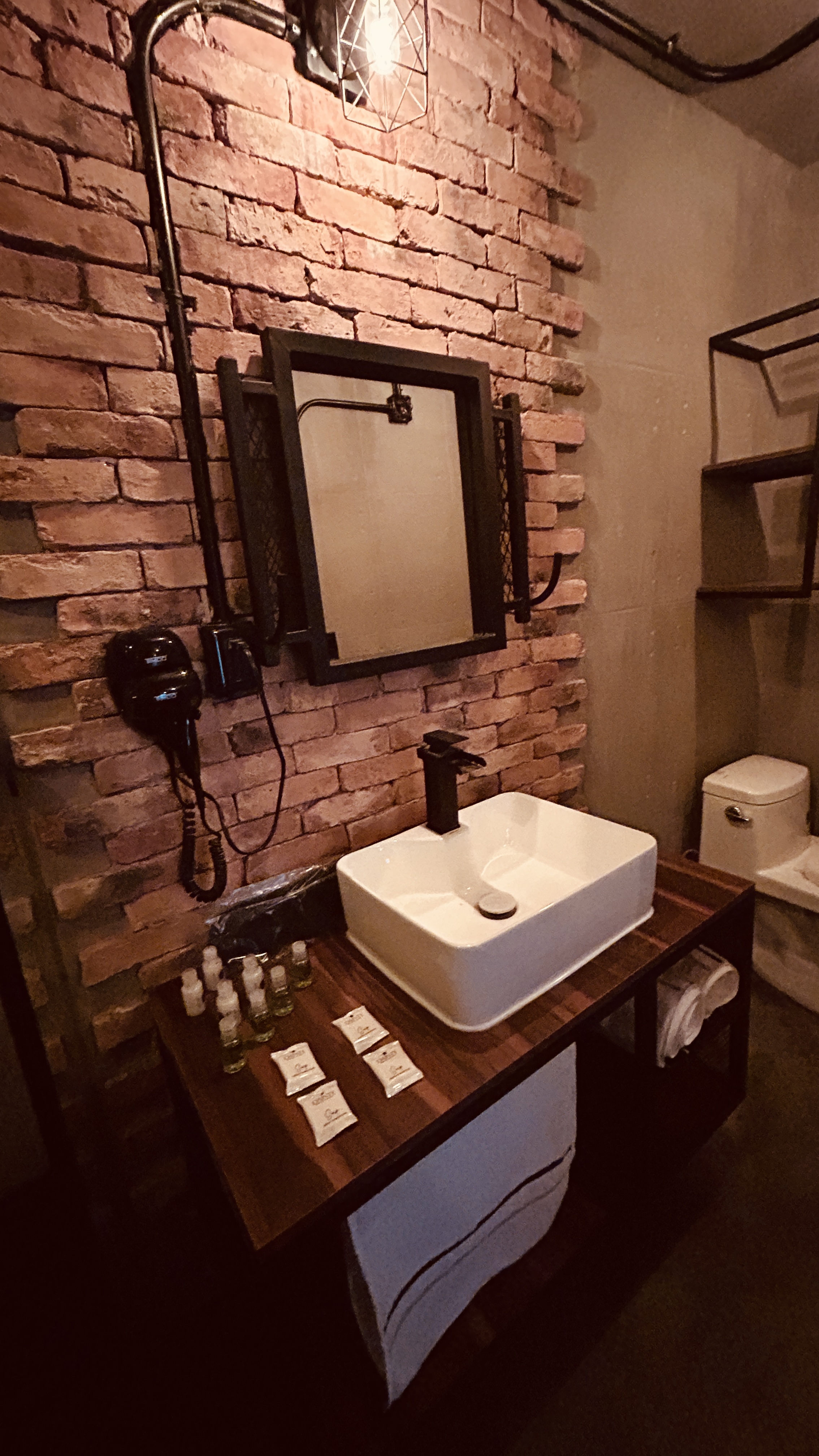 Baño industrial con muro de ladrillo expuesto, lavabo blanco y grifería negra en Hotel Van Gogh